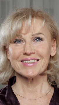 Close up portrait of an attractive mature blonde woman with blue eyes looking at the camera, initially with a neutral expression, and then slowly breaking into a warm and beautiful smile
