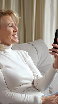 Smiling mature blonde woman relaxing on a comfortable sofa at home while making a video call, waving her hand and gesturing happily to her family or friends on the smartphone screen