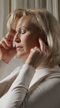 Mature blonde woman suffering from a terrible headache, massaging her temples and neck in pain while feeling unwell, sick, stressed, and tired, sitting at home in a dimly lit room