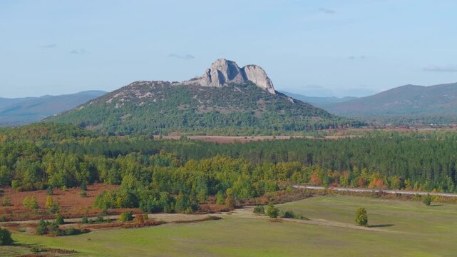 AERIAL: Scenic rocky mountain rising above forests, green fields and colourful autumn vegetation near Otočac, Croatia. Clear blue sky and layered hills create a broad, picturesque rural landscape.