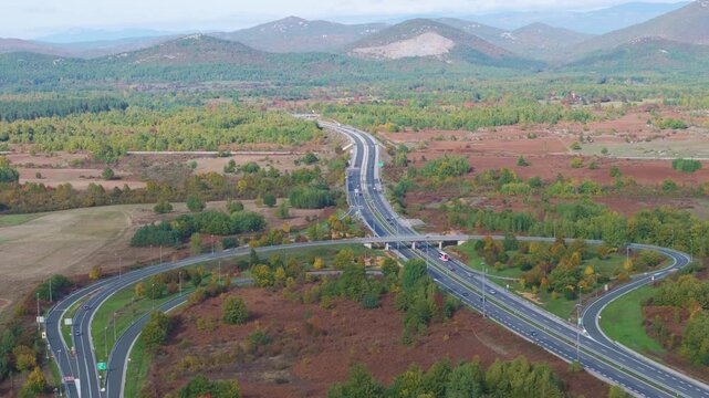 AERIAL: Large highway interchange surrounded by fields and colorful autumn woodland in Croatia. Curving ramps, bridges and moving vehicles cross a broad rural valley beneath layered mountain ridges.
