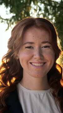 Close up portrait of a charming young woman with red hair and freckles smiling sincerely at the camera while standing in a park. Her hair beautifully illuminated by the warm light of the setting sun