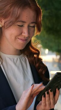 Professional woman with red hair and freckles happily scrolling through her smartphone's social media feed, enjoying her online communication and networking in a sunlit park outdoors