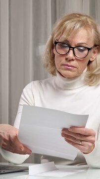Stressed senior woman with glasses sitting at a desk opening a letter and becoming upset while reading the bad news, showing expressions of sadness, concern, and disappointment