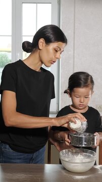 Loving mother and her cute little daughter sifting flour together into a glass bowl while cooking in the kitchen, enjoying a bonding moment and developing skills through a family activity