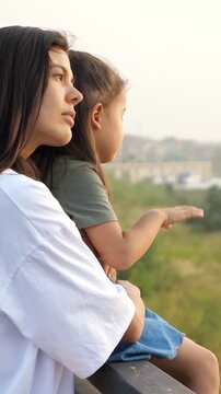 Loving young mother embracing her little daughter while they stand on a bridge, enjoying the scenic view and spending quality time together outdoors during a beautiful summer evening