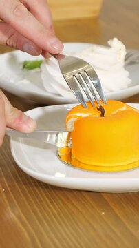 Close up view of a person using a knife and fork to cut a slice from an orange mousse cake shaped like an apple, revealing the jelly filling inside on a white plate in a cozy cafe setting