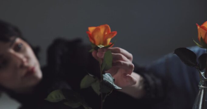Woman touches a single vibrant orange rose while resting her hand near the stem, showing soft focus on her dark hair and a neutral gray background which provides elegant artistic mood and composition.