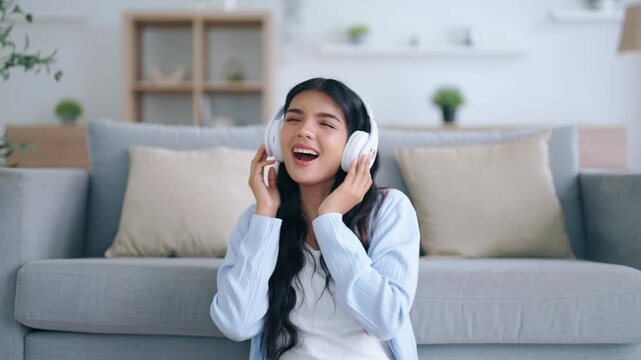Young asian woman listening to music on couch in living room at home. Happy asia female wearing headset and sitting on the floor