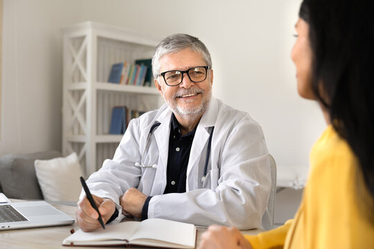 Smiling senior male doctor sit at desk in clinic, making notes, writing information, listening female patient during medical consultation. Professional healthcare services provider portrait concept