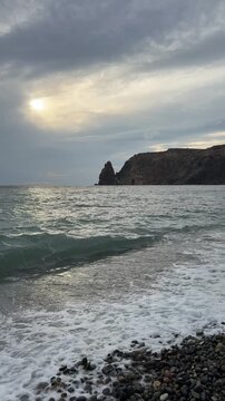 Coastal ocean waves crashing onto a pebbled beach shore with rocky cliffs at sunset. Static camera shot.