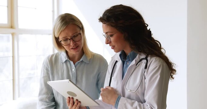 Positive young physician woman explaining diagnosis to female patient, showing screening result on tablet, speaking, giving instructions for takin meds, recommendation, consultation