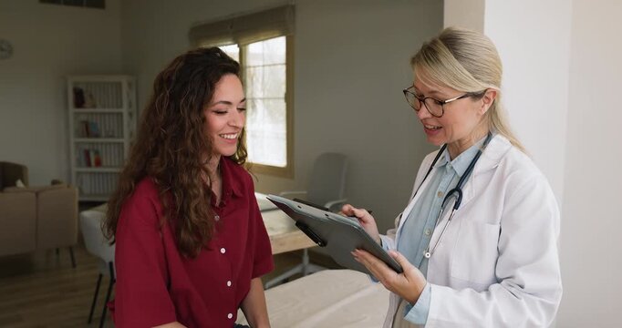 Positive friendly doctor woman talking to young female patient about healthcare, health complaints in medical examination room, filling up form for records, holding clipboard, writing, smiling