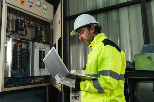 Professional electrical technician using laptop checking electrical system inspection beside control panel in factory. diagnostics, troubleshooting, safety, preventive service.