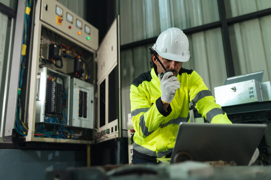 Professional electrical technician communicates by walkie talkie while using laptop checking electrical system inspection beside control panel. diagnostics, safety, preventive maintenance.