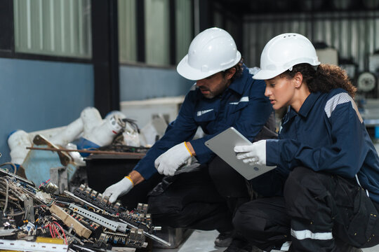 Environmental engineers sort electronic components and industrial scrap beside robotic machinery, using tablet data for waste management, responsible disposal, sustainability management.