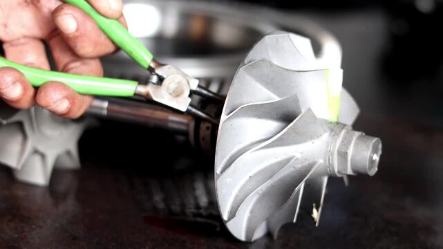 Technician handling and servicing metal turbocharger turbine compressor wheel with green handled pliers in workshop.