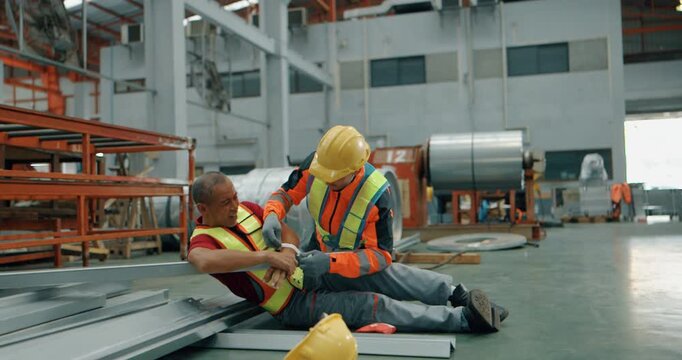 Injured factory worker in pain on floor after accident while colleague helps in steel warehouse
