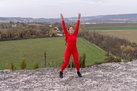 Energetic woman jumping in red sportswear on rocky cliff overlooking rural landscape