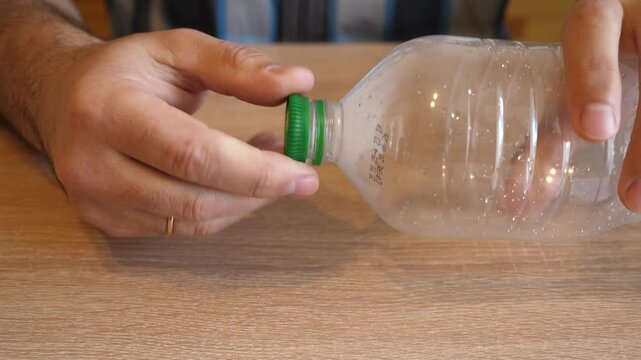 Person unscrewing and screwing the cap of an empty plastic bottle in close up on a wooden table. Continuous action with original sound.