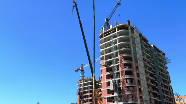Residential high-rise construction site viewed from ground level with elevator going down, multiple cranes and blue sky