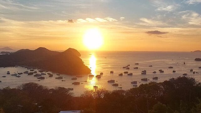 An elevated view revealing a stunning golden sunset over the tropical waters of Labuan Bajo, East Nusa Tenggara, Indonesia. The warm sunlight creates a brilliant reflection on the water's surface, ill