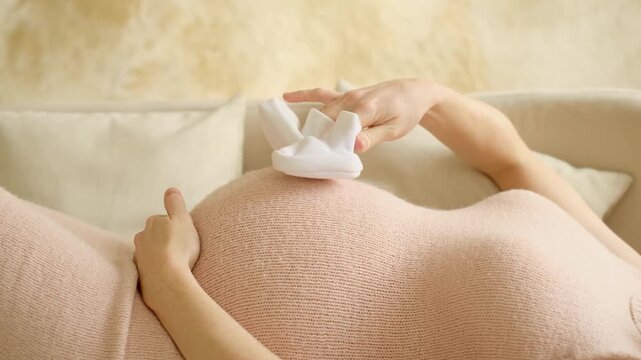 a pregnant woman pats her stomach while holding baby booties or socks, a woman waiting for a baby to be born, close-up of a pregnant woman's stomach