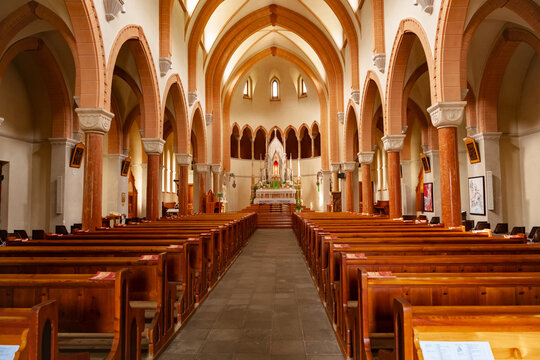 Interior view of the Church of San Sebastiano in Falcade Alto, showing a symmetrical nave with rows of wooden pews, warm-toned columns, pointed arches. Dolomites, Italy