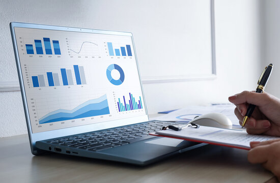 Business analyst working on laptop analyzing financial dashboard graphics and writing notes on document clipboard in office desk.
