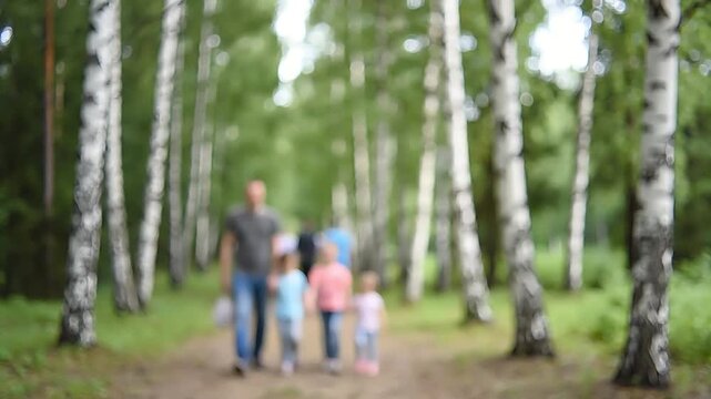 Blurred image of a family, a man and three children, walking together on a dirt path through a birch forest on a sunny day.