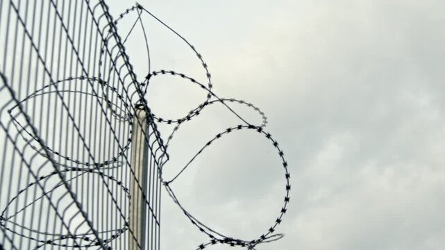 Close-up video of razor wire mounted on a tall metal security fence against an overcast sky. Symbolic security, restricted access, border protection, prison perimeter, surveillance, safety.