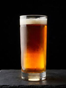 Full pint glass of chilled golden amber craft beer with thick white foam head isolated on dark studio background refreshment beverage concept
