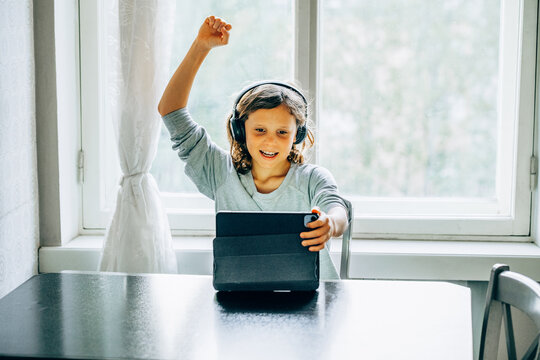 Happy long haired child boy  celebrating success while using tablet for online learning at home.