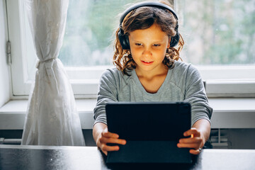 Focused long haired child boy using digital tablet with headphones during online learning at home. © Maria