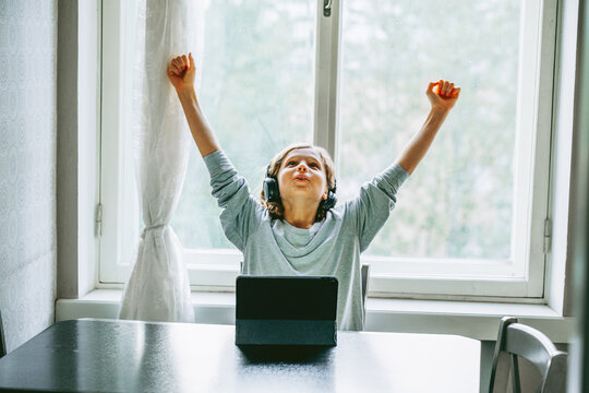 Young child wearing headphones raises both arms in celebration after achieving success on a digital tablet. Bright natural light and a comfortable home setting illustrate online learning.