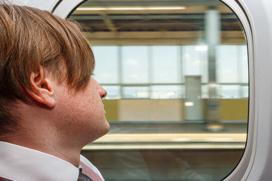 Close-up profile of a young businessman looking out a high-speed train window as a modern railway station platform blurs past.