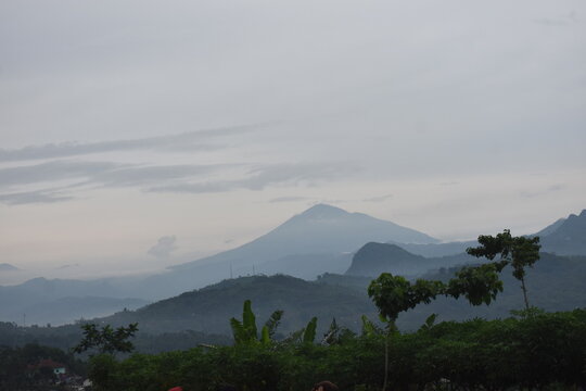 Distant mountain peaks rise above misty, layered hills. Green forests blanket rolling terrain below. Soft clouds hover over the distant summit. Villages nestle quietly in the valley's embrace