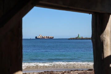 Obraz premium Perth, Western Australia, Australia - July 30 2026: MSC container ship, boat entering Fremantle harbor. Seen from Bathers Beach, framed by wooden timbers. South mole green lighthouse