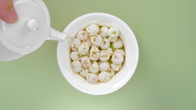 Overhead macro video of hot water pouring from white teapot into ceramic cup with fresh white jasmine buds and dried tea leaves, traditional chinese tea brewing ceremony background for commercial use