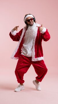 Trendy man in Santa costume dancing with headphones on pink background