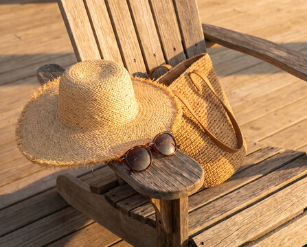 Wide-brimmed straw sun hat and sunglasses staged beside a raffia tote on a wooden beach-chair armrest