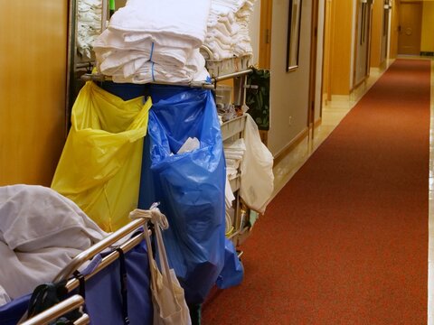 A hotel housekeeping cart loaded with folded white linens, towels, and laundry bags stands in hallway. Bright yellow and blue cleaning bags hang from the side, depicting daily room service