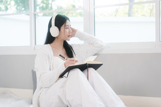 Young woman in white loungewear sits by a bright window, wearing headphones and writing in a notebook. Calm home scene with soft light, focus, study, journaling, and relaxation.