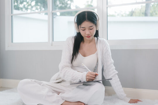 Young woman in white loungewear sits by a bright window, wearing headphones and writing in a notebook. Calm home scene with soft light, focus, study, journaling, and relaxation.