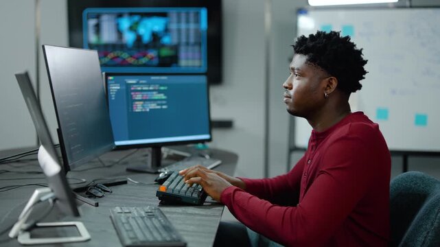 Focused Young African American software developer coding on multiple monitors in a modern tech office. IT professional analyzing complex data charts and graphs. Skilled cybersecurity expert typing.