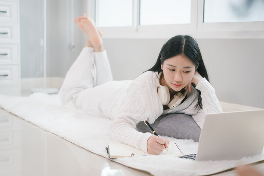 Young woman in a white sweater lying on a bed, writing in a notebook beside a laptop and headphones in a bright cozy room, showing study, remote work, and relaxed home life.