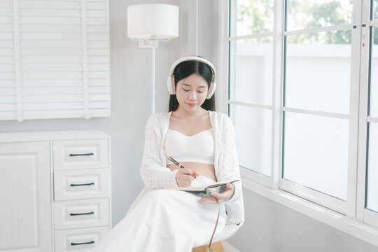 Young woman in white casual clothes using a smartphone and wearing headphones by a bright window at home, enjoying music and a relaxed morning mood.
