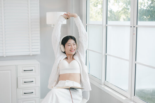 Young woman in white casual clothes using a smartphone and wearing headphones by a bright window at home, enjoying music and a relaxed morning mood.
