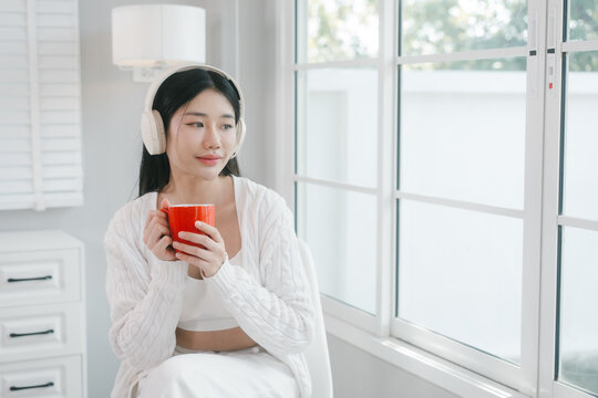 Young woman in white loungewear sits by a bright window, wearing headphones and drinking from a red mug in a calm modern home setting