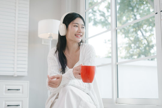 Young woman in white loungewear sits by a bright window, wearing headphones and drinking from a red mug in a calm modern home setting